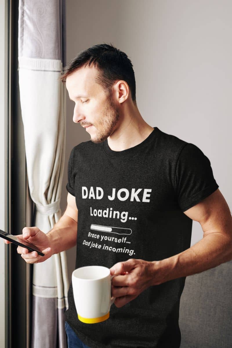funny dad shirt with a man wearing a black t-shirt with text 'Dad Joke Loading, Brace yourself…Dad joke incoming.', holding a mug and phone.