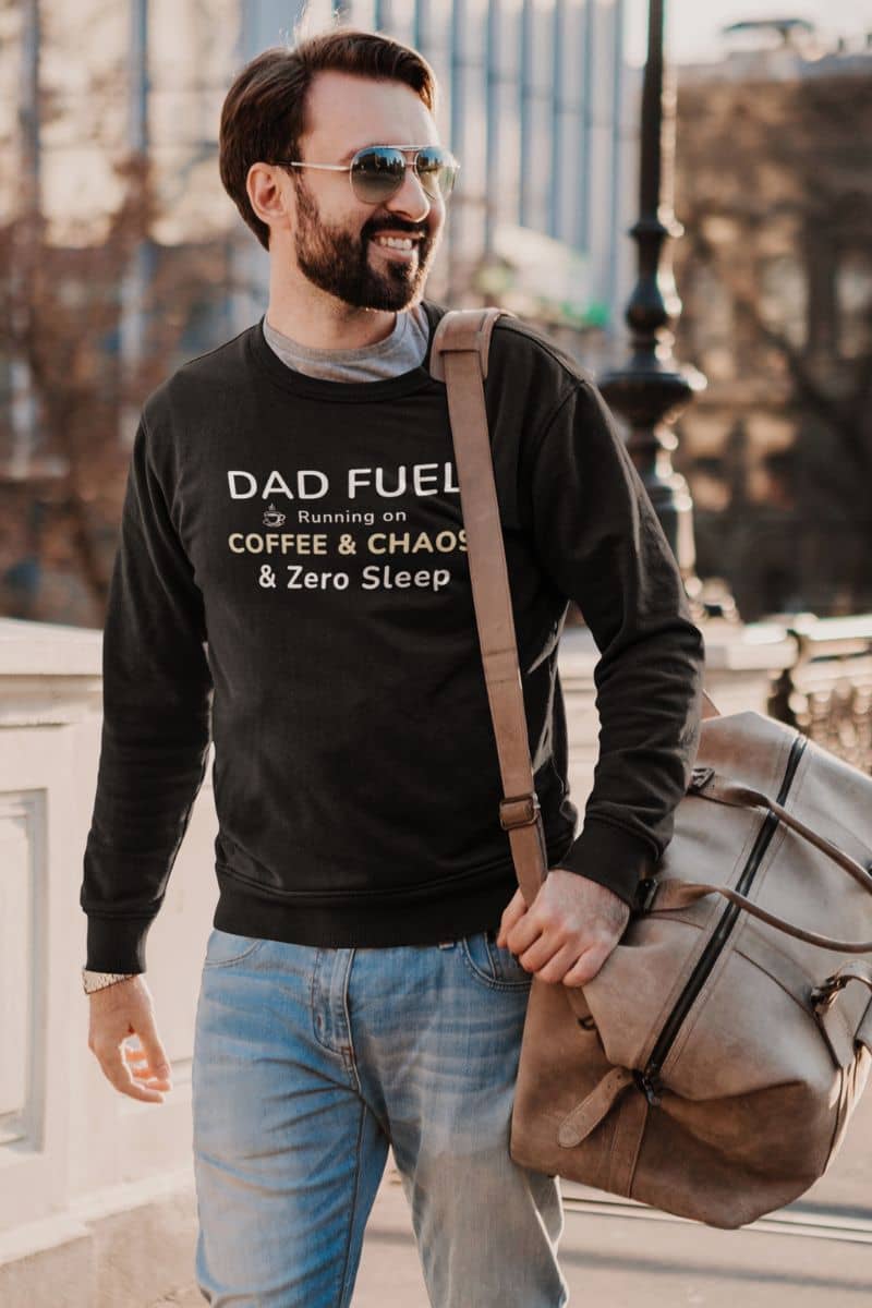 funny dad sweatshirt with a man wearing a black sweatshirt with text 'DAD FUEL – Running on Coffee & Chaos & Zero Sleep', holding a brown bag in an urban setting.