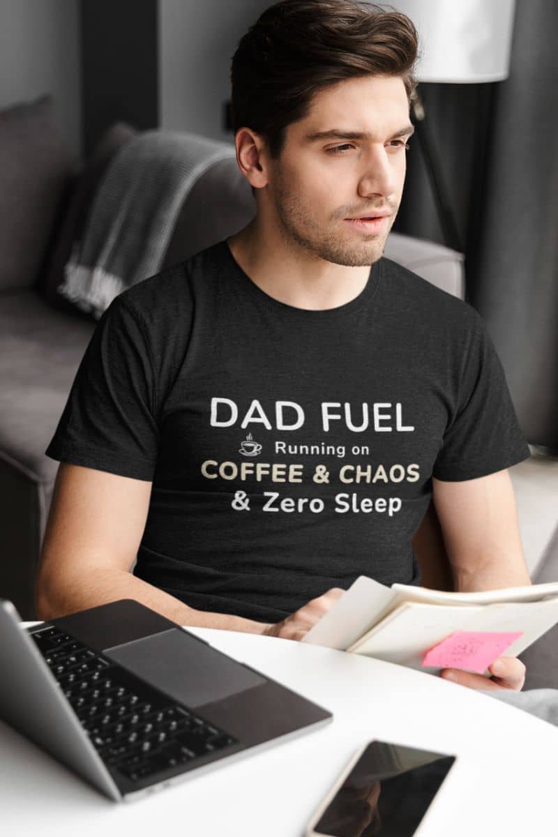 funny dad tshirt with a man wearing a black t-shirt with text 'DAD FUEL – Running on Coffee & Chaos & Zero Sleep', sitting at a desk with a laptop and tablet.