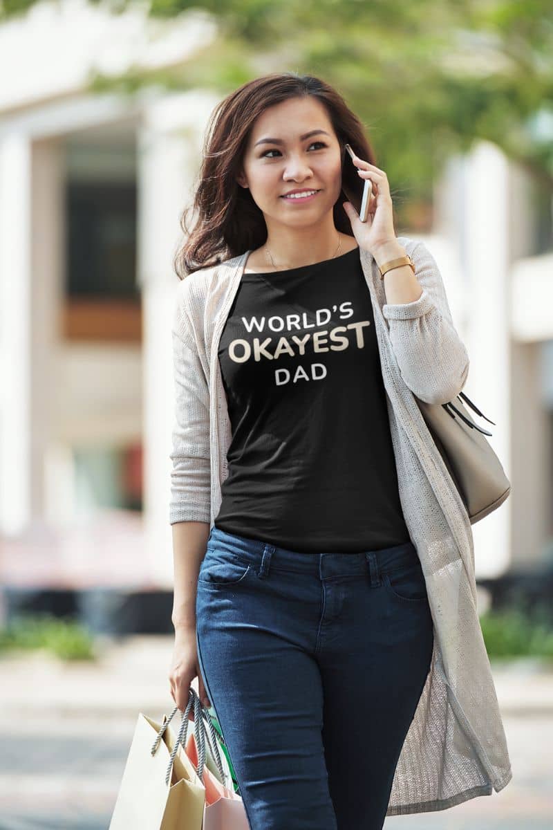 funny father shirt with a woman wearing a black t-shirt with 'World's Okayest Dad' text, standing outdoors.