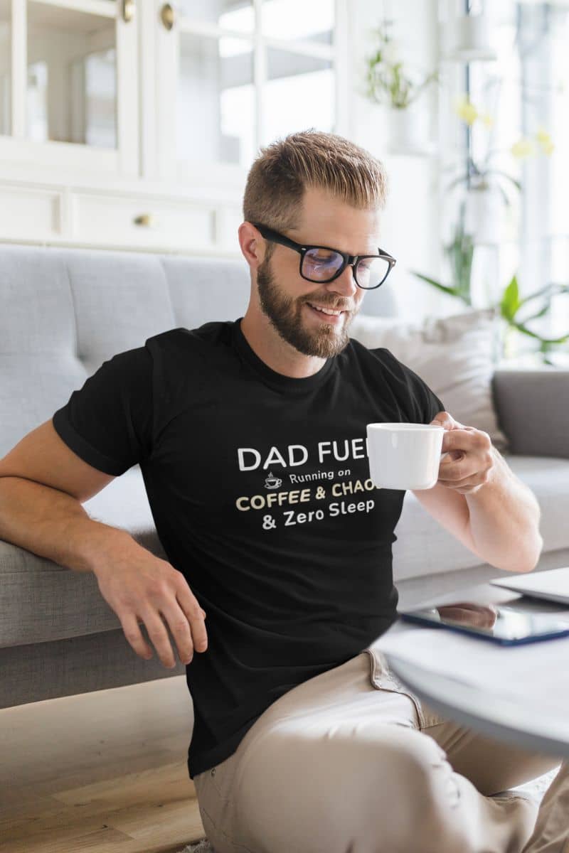 funny father t shirts with a man sitting on a couch holding a coffee cup, wearing a black t-shirt featuring the text 'DAD FUEL – Running on Coffee & Chaos & Zero Sleep'