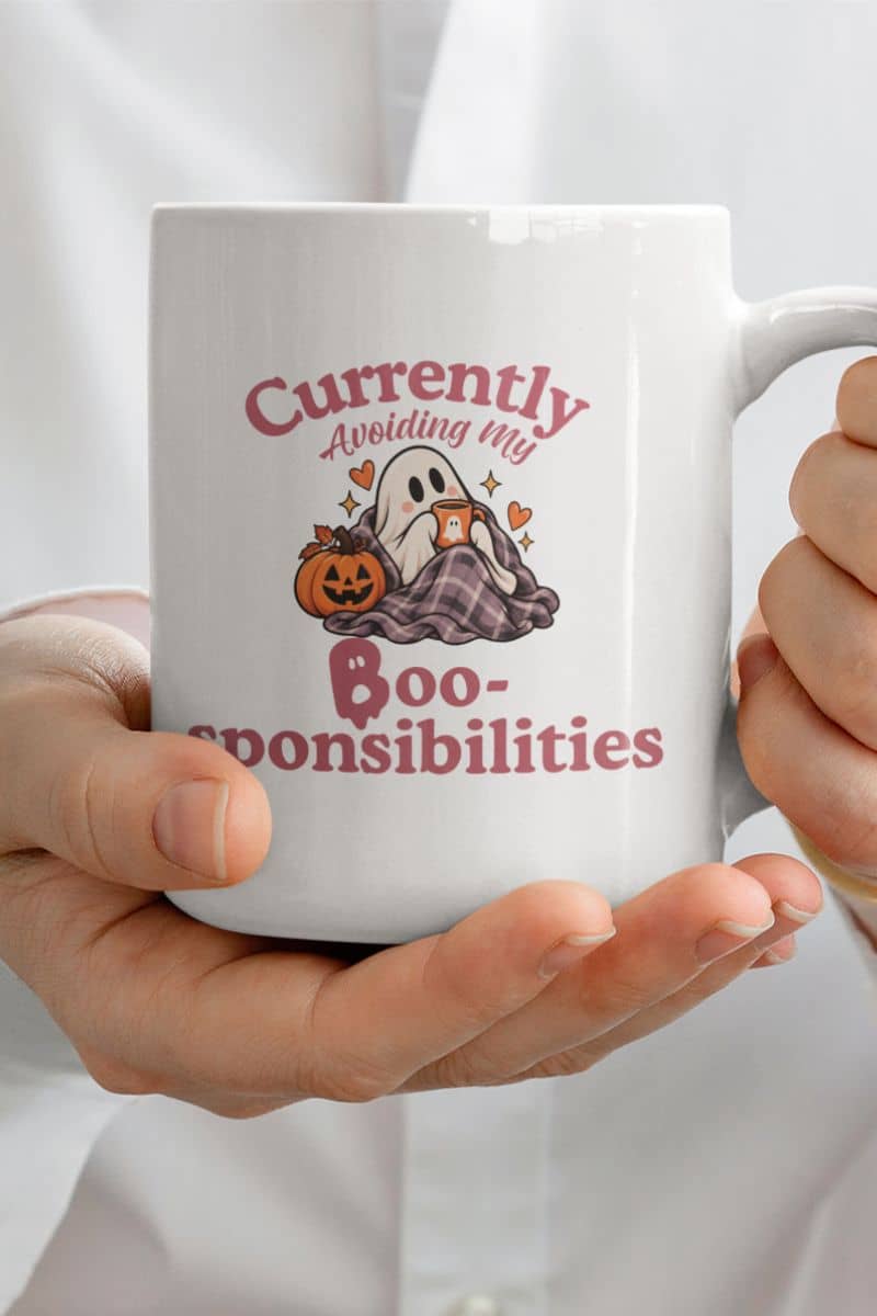 A person holding a funny ghost mug with a Halloween-themed graphic that reads 'Currently Avoiding My Boo-sponsibilities' in pink text, featuring a ghost, pumpkin, and plaid blanket.