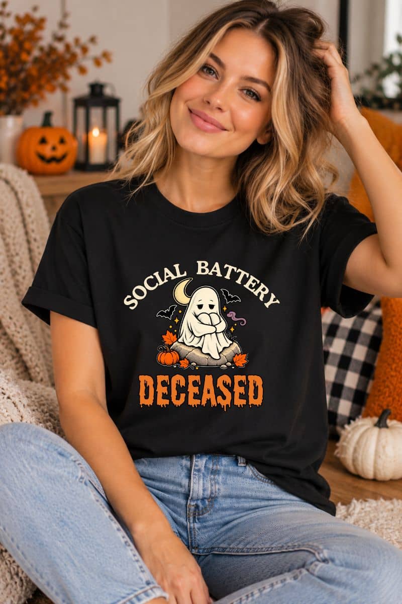 A woman wearing a funny ghost shirt featuring a 'Social Battery: Deceased' graphic with a ghost and pumpkins, sitting in a cozy, decorated home setting.