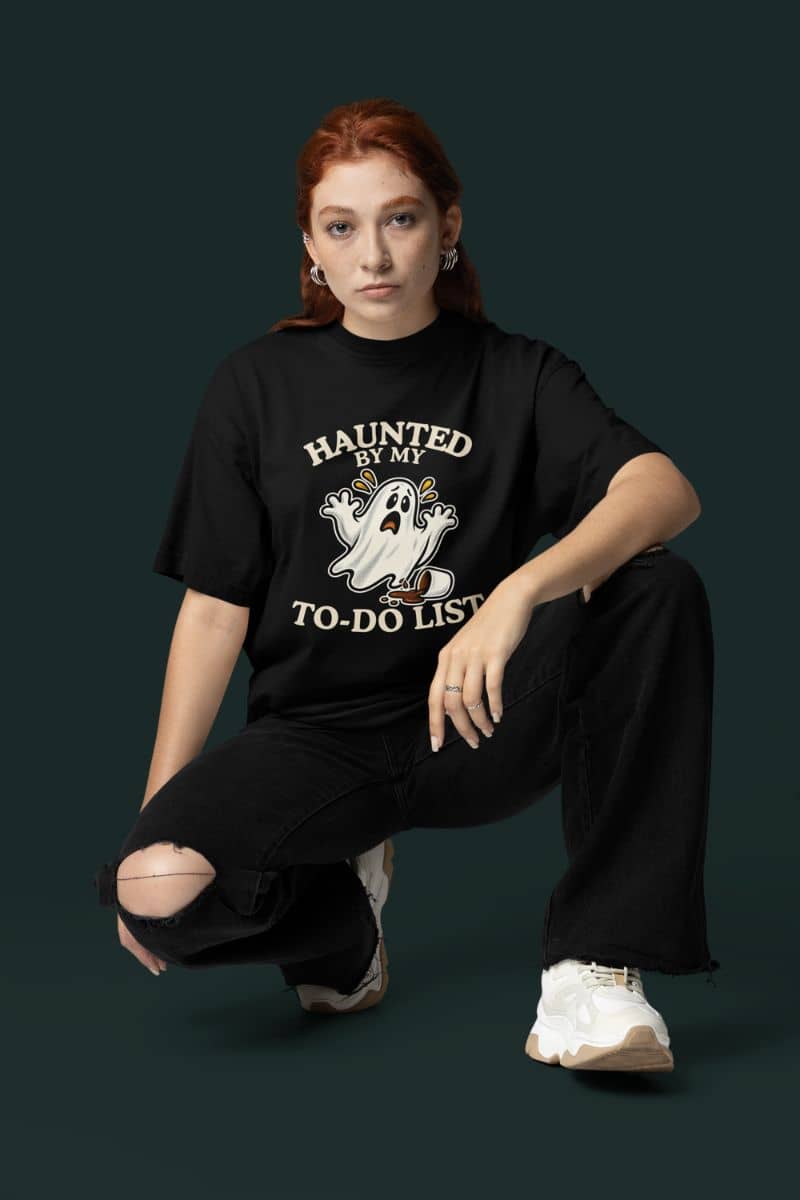 A woman wearing a funny ghost shirt featuring a “Haunted by My To-Do List” funny ghost, posting in front of a dark green background