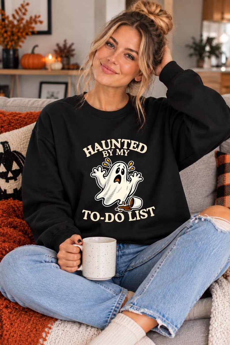 A woman wearing a funny ghost sweatshirt featuring a “Haunted by My To-Do List” funny ghost, sitting in the living room