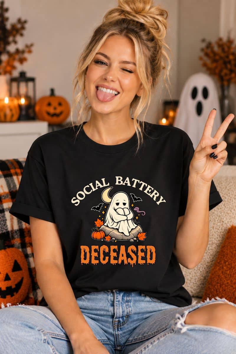 A woman wearing a funny Halloween t shirts featuring a 'Social Battery: Deceased' graphic with a ghost and pumpkins, sitting in a cozy, decorated living room.