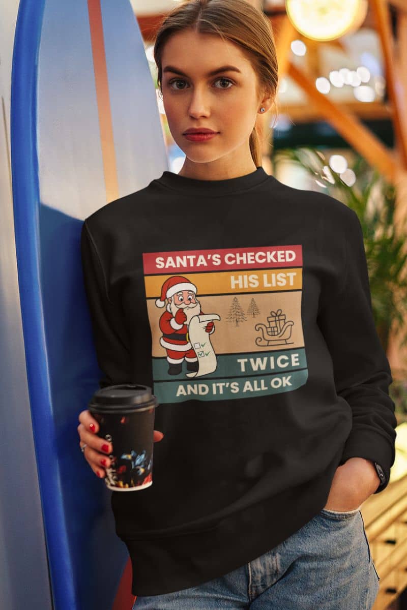 funny holiday sweatshirt with a woman wearing a black sweater with the quote 'Santa's Checked His List Twice And It's All Ok', holding a coffee cup.