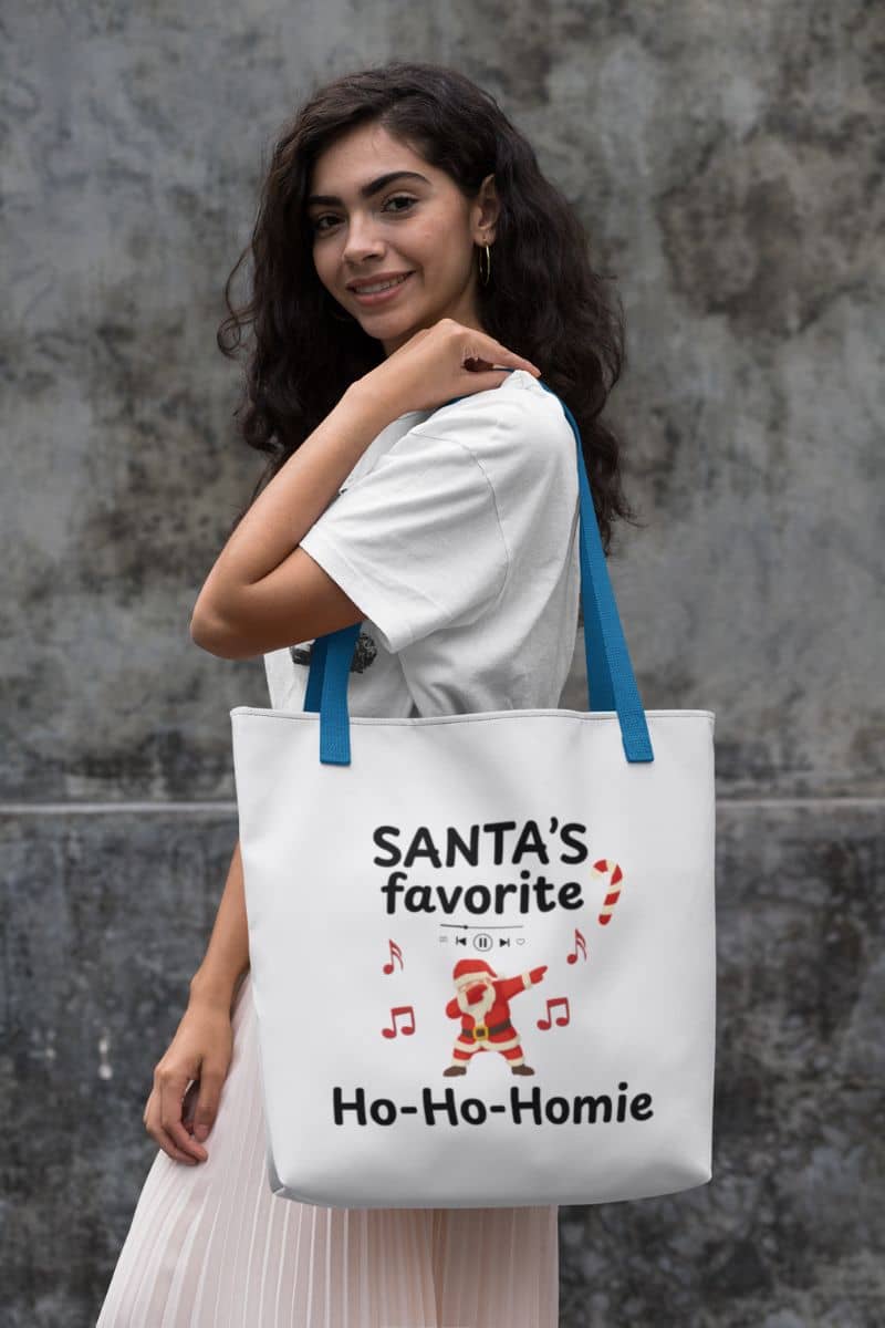 funny holiday tote with a woman holding a tote bag with text 'Santa's Favorite Ho-Ho-Homie' against a gray wall.