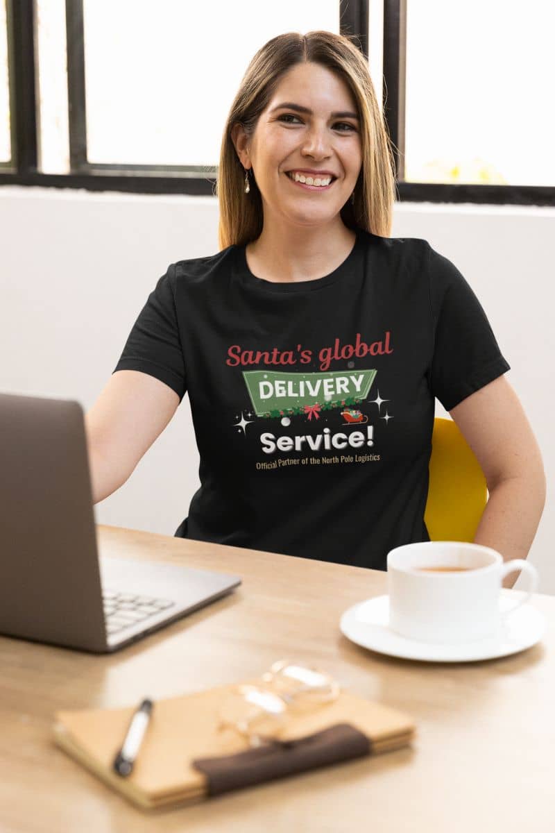 funny holiday t-shirt with a woman wearing a black t-shirt with text 'Santa's Global Delivery Service.', sitting at a desk with a laptop and coffee.