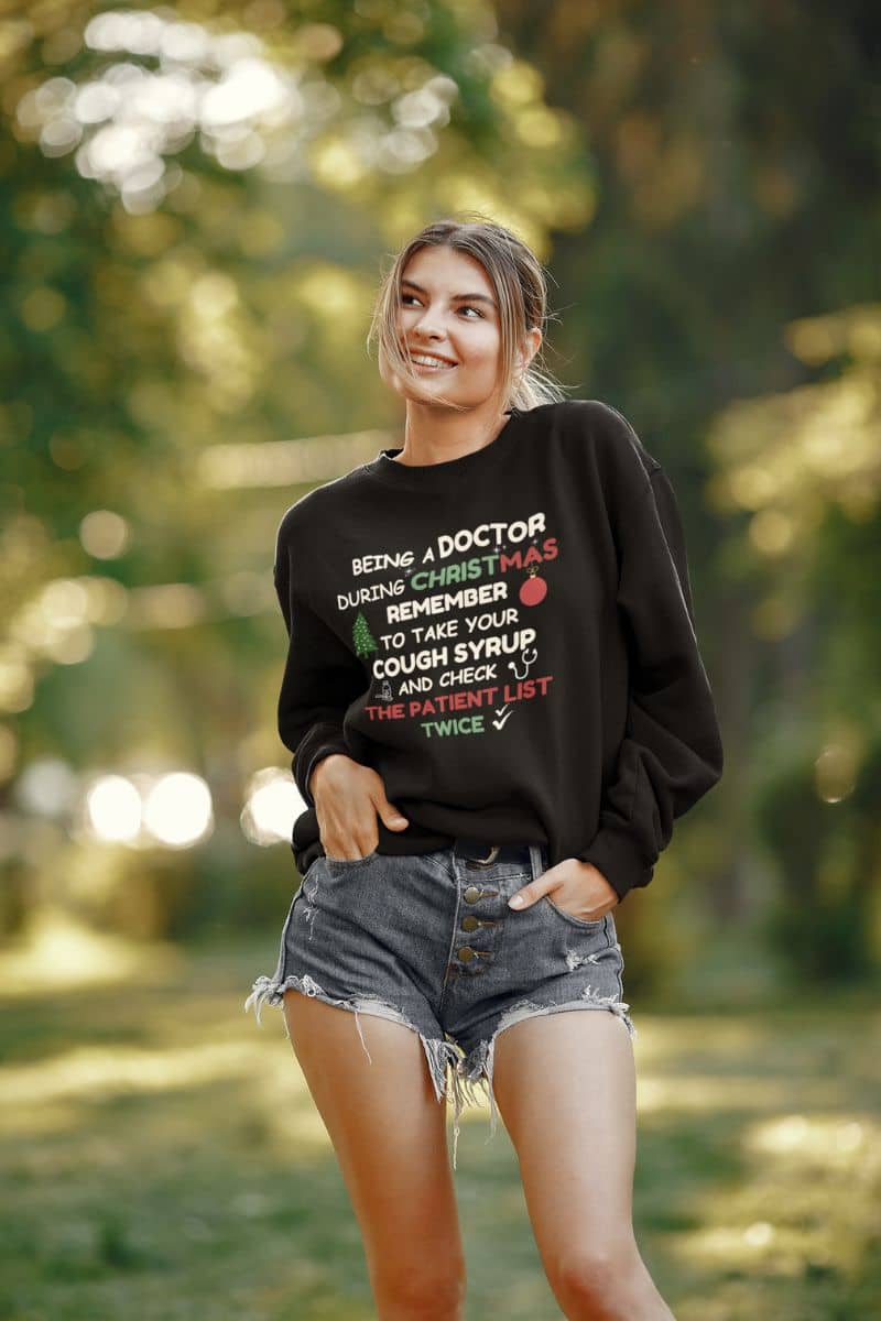 funny hospital christmas pullover with a woman wearing a black sweatshirt with text 'Being a doctor during Christmas, remember to take your cough syrup and check the patient list twice' in a forest setting