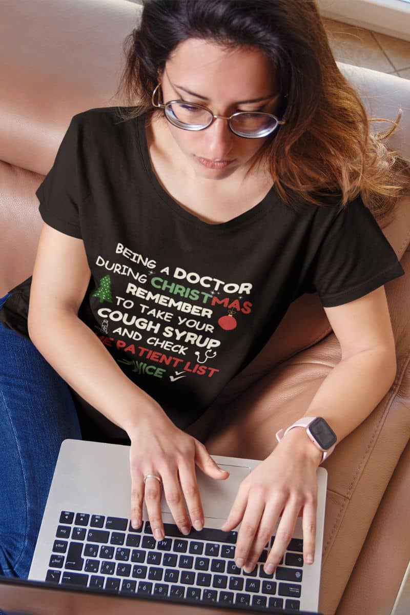 funny medical christmas shirt with a woman wearing a black t-shirt with a humorous Christmas doctor's office message, using a laptop.