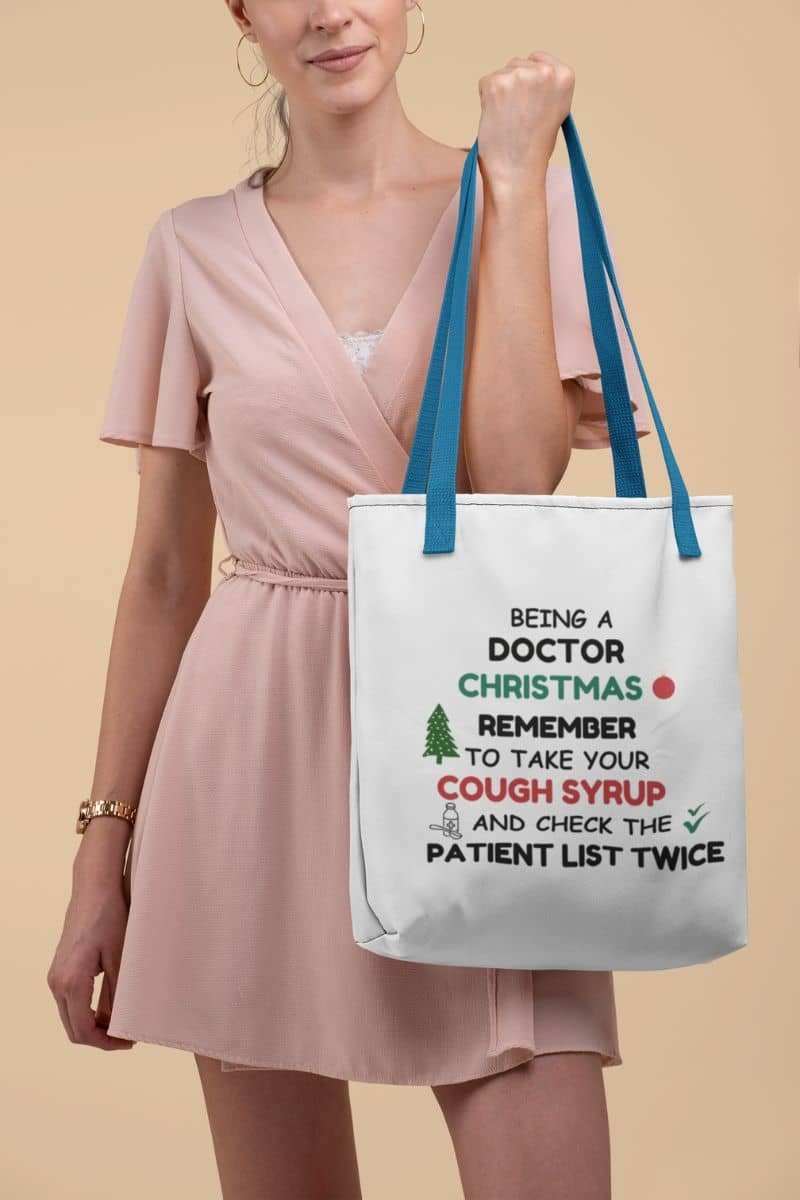 funny medical tote bag with a woman holding a tote bag featuring the text 'Being a doctor during Christmas, remember to take your cough syrup and check the patient list twice' on a beige background