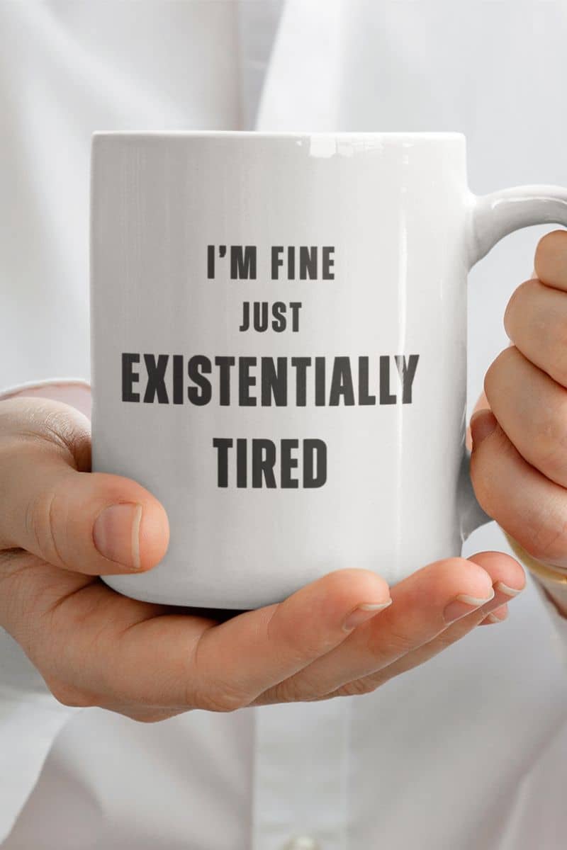 funny mental health mug with a person holding a white mug with text 'I'm fine just existentially tired' against a light background