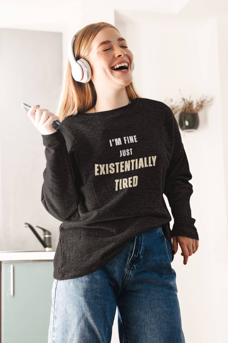 funny mental health sweatshirt with a person wearing a black sweatshirt with text 'I’m fine. Just existentially tired.', laughing in a kitchen.