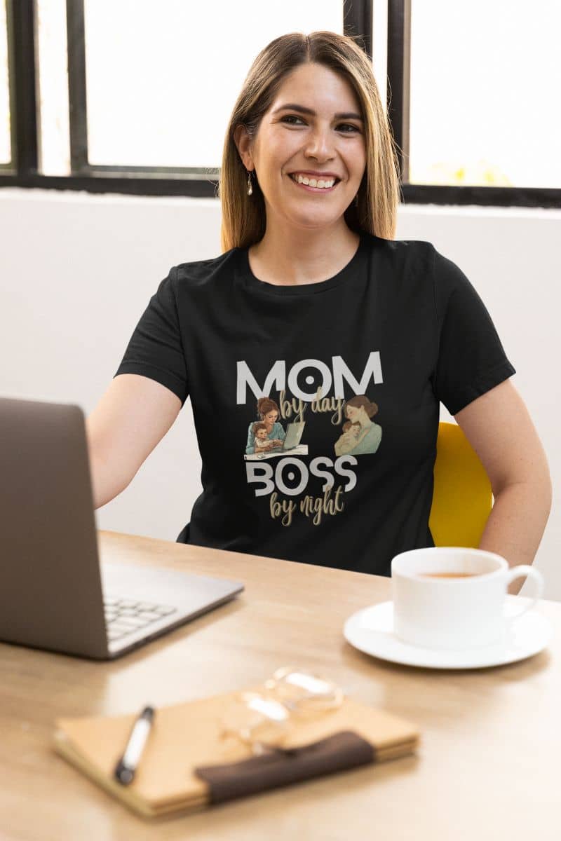 funny mom graphic tee with a woman wearing a black t-shirt with 'Mom by day, Boss by night' text, sitting at a desk with a laptop and coffee.