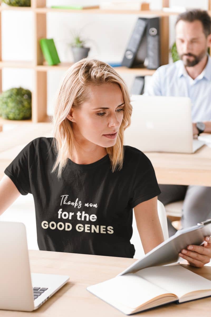 funny mom graphic tee with a woman in a black t-shirt with text sitting at a desk with a laptop and book, with a man in the background.