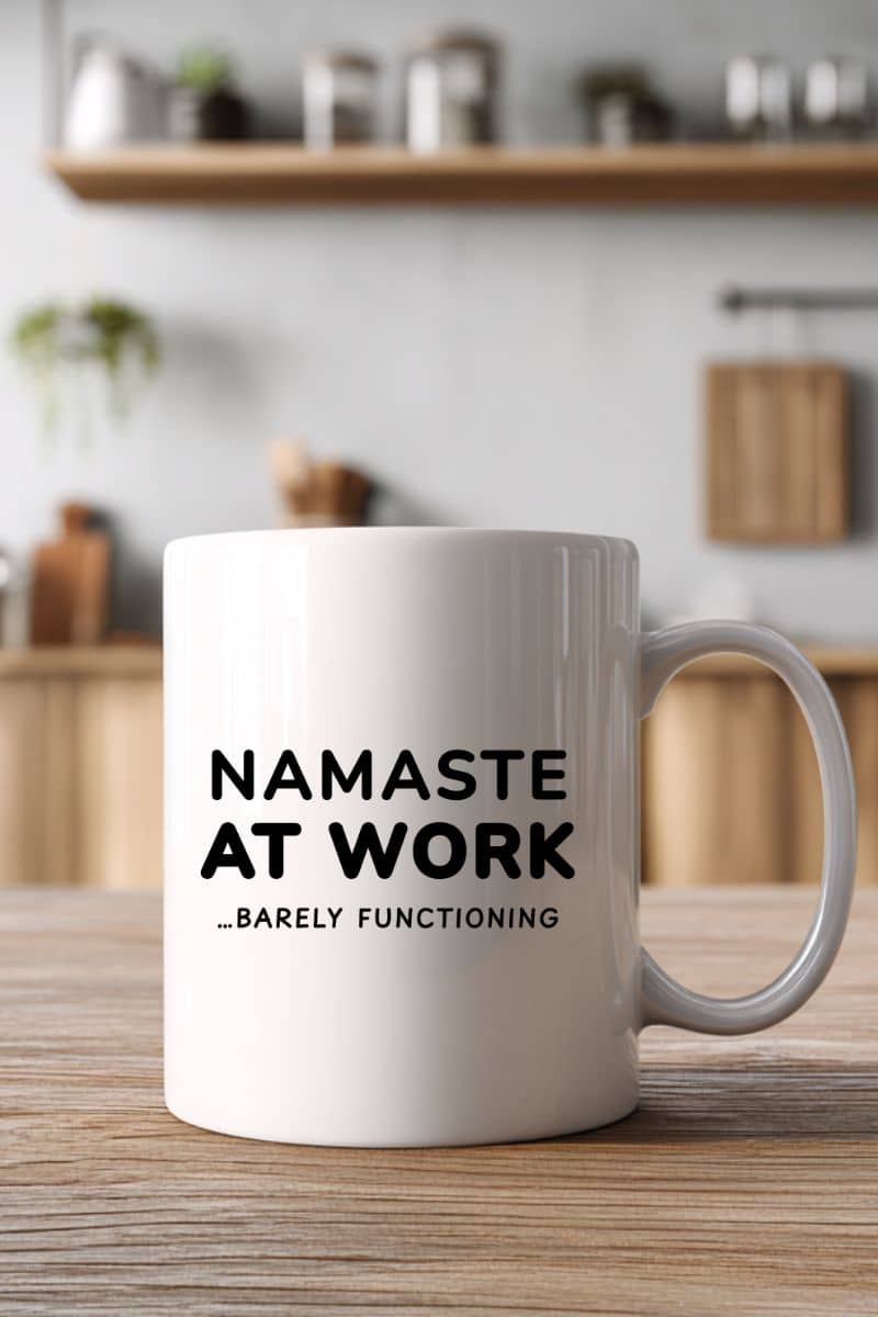 funny mug for colleague with a white mug with text 'Namaste at work... barely functioning' on a wooden table in a kitchen setting.