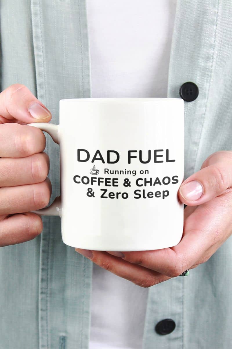 funny mugs for dads featuring a person holding a mug with text 'DAD FUEL – Running on Coffee & Chaos & Zero Sleep', wearing a light gray shirt.