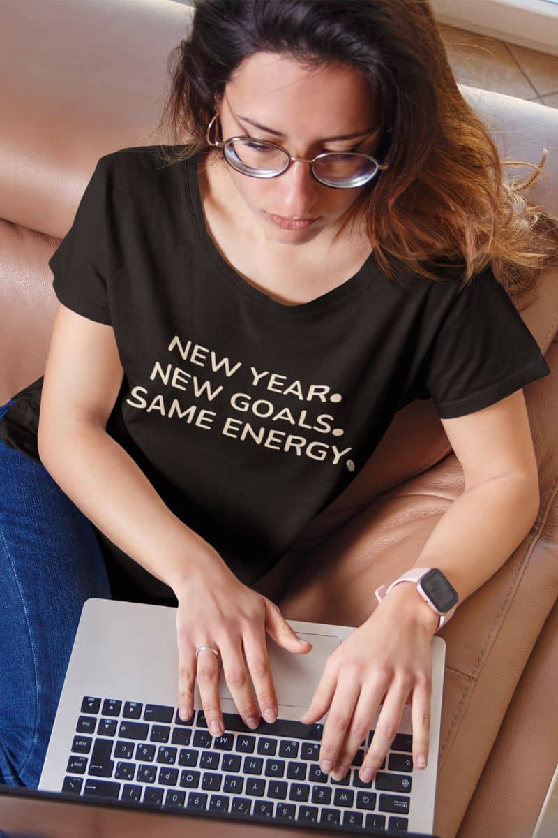 funny new year t shirt with a woman wearing a black t-shirt with motivational text 'New Year. New Goals. Same Energy.', using a laptop.