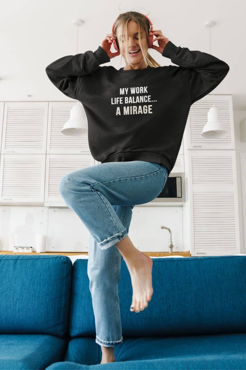 funny office apparel with a person wearing a black sweatshirt with text 'My Work-Life Balance Is a Mirage', standing on a blue couch in a kitchen.