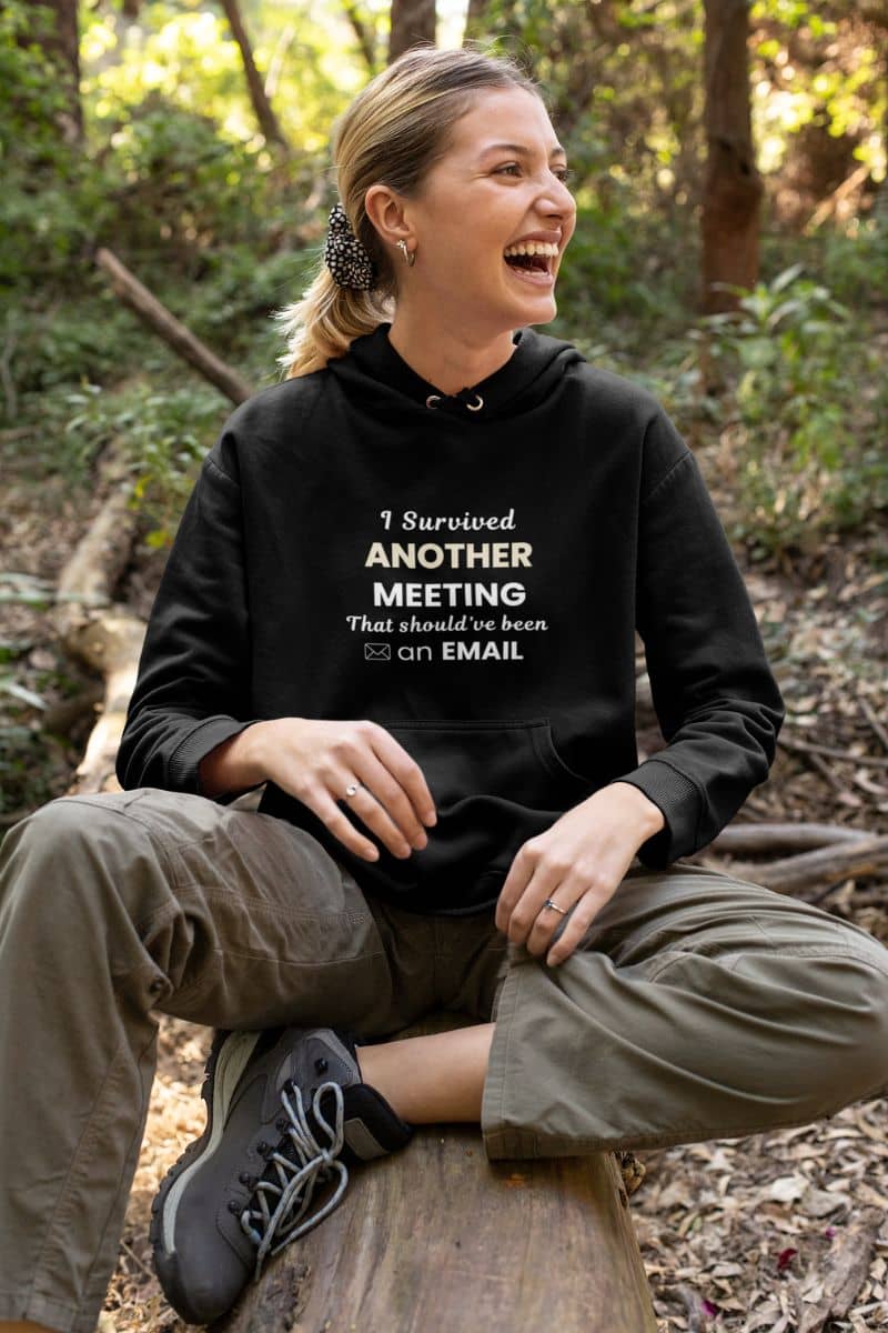 funny office hoodie with a person wearing a black hoodie with text  'I Survived Another Meeting That Should’ve Been an Email' in a forest setting