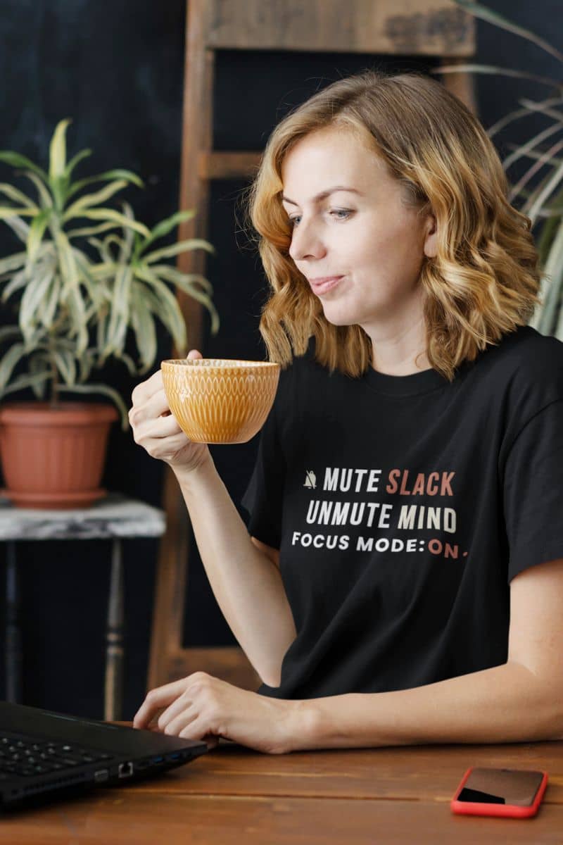 funny office stress shirt with a woman holding a mug with text on her shirt, sitting at a desk with a laptop and phone.