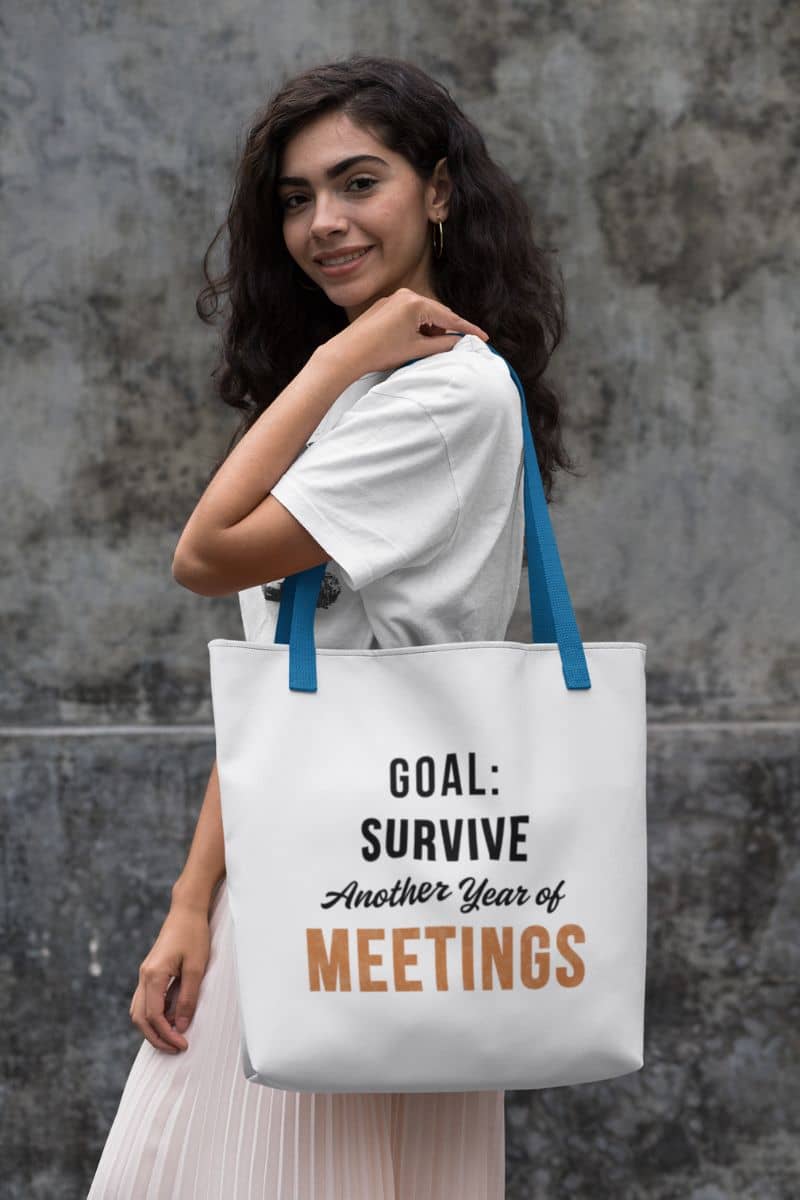 funny office tote bag with a woman holding a tote bag with text 'Goal: Survive Another Year of Meetings' against a gray wall.