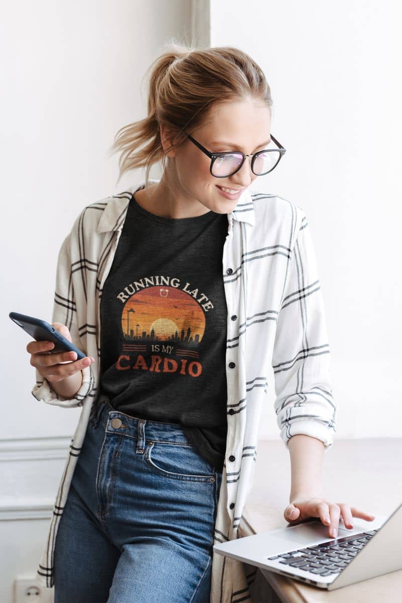 funny quote shirt with a woman wearing a black t-shirt with 'Running Late Is My Cardio' design, using a laptop.