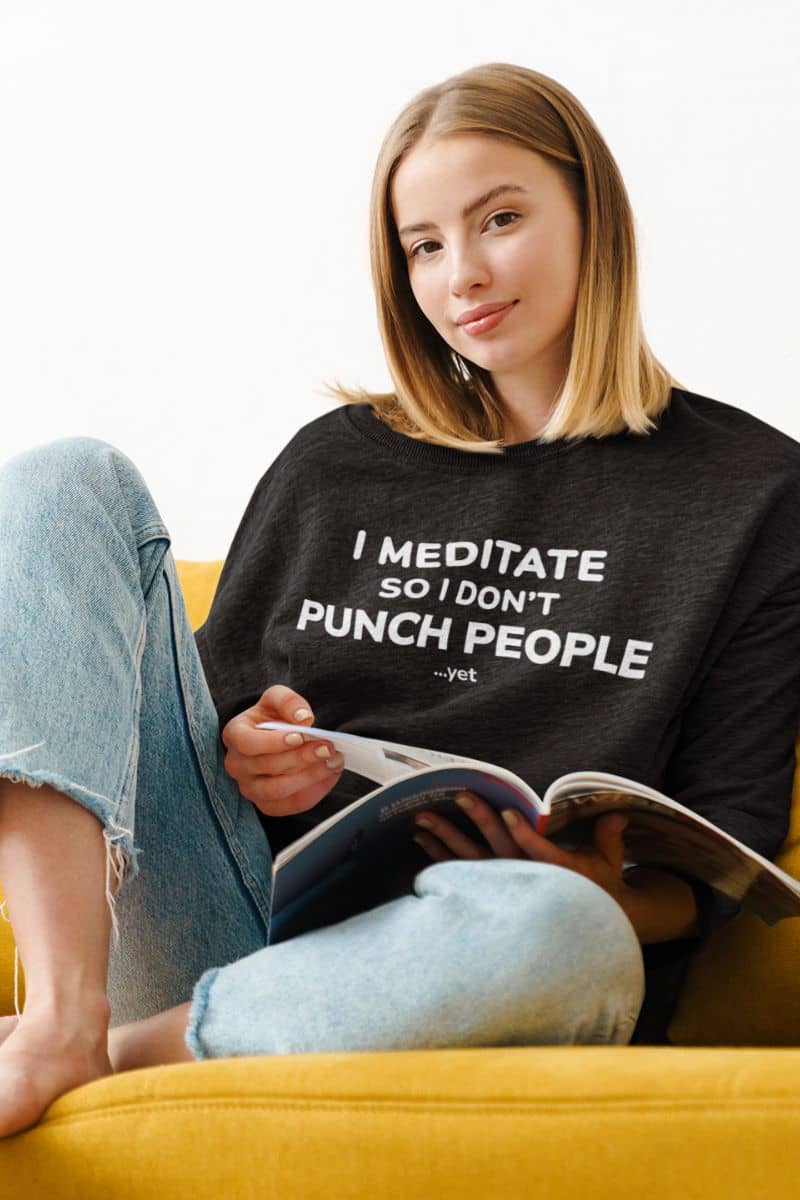 funny quote sweatshirt with a woman wearing a black sweatshirt with text 'I Meditate So I Don’t Punch People...yet', sitting on a yellow couch reading a book.