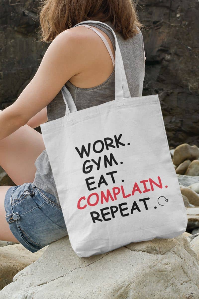 funny reusable shopper with a person holding a tote bag with text 'Work. Gym. Eat. Complain. Repeat.' on a rocky surface.