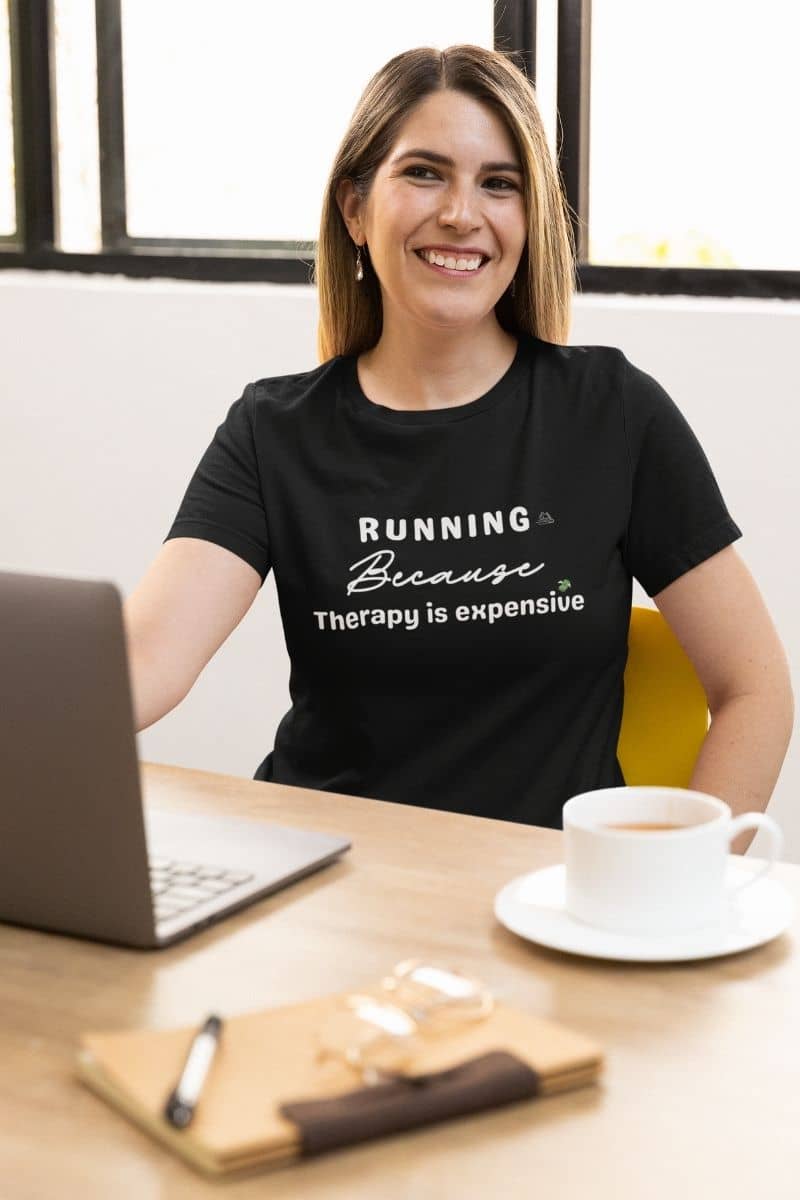 funny runner tee worn by a woman sitting at her desk, featuring the text 'Running because therapy is expensive'