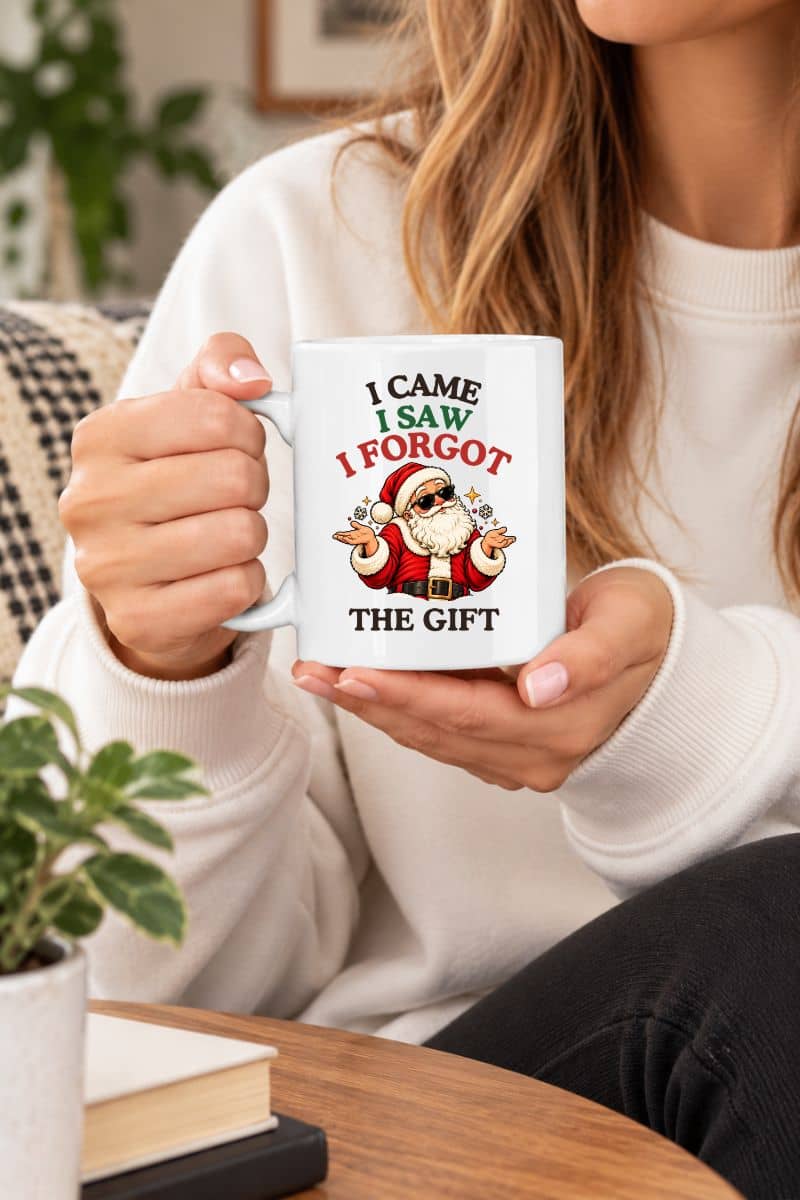 Woman in a white sweater holding a funny Santa mug with a shrugging Santa and “I Came. I Saw. I Forgot the Gift” graphic.