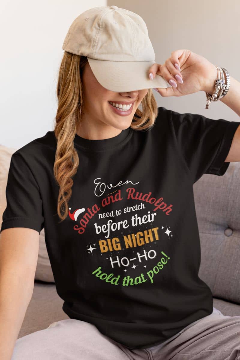 funny santa yoga shirt with a person wearing a black t-shirt with text 'Event Santa & Rudolph Need to Stretch Before their Big Night. Ho-Ho-Hold That Pose!', sitting on a couch.