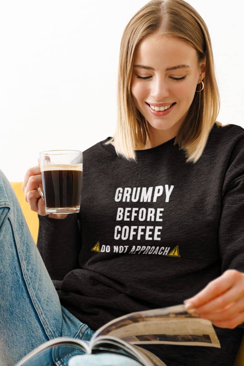 funny sarcastic sweatshirt with a woman wearing a black sweatshirt with 'Grumpy before coffee, do not approach text, holding a coffee cup and reading a newspaper.