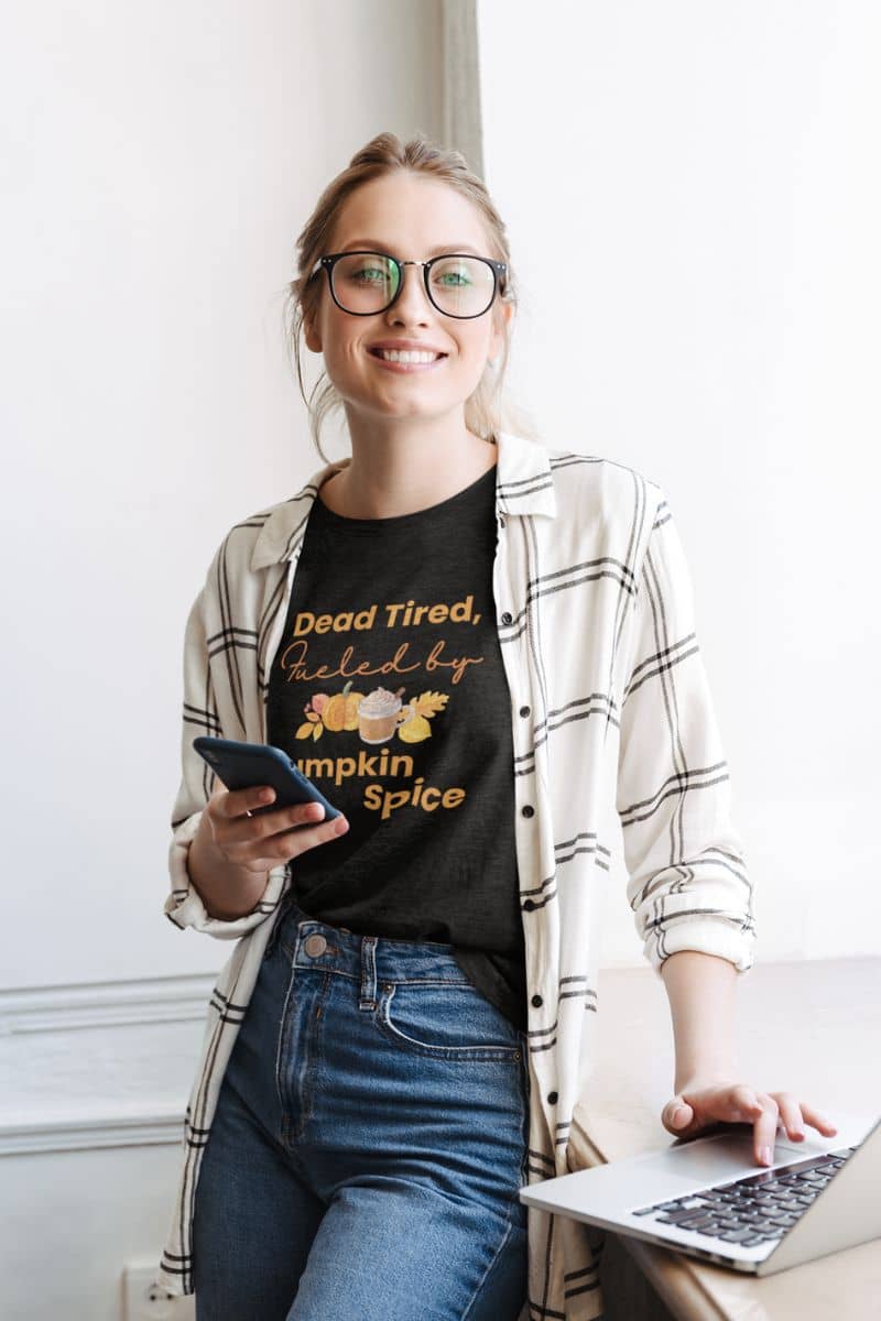 funny seasonal tee with a woman wearing a black t-shirt with text 'Dead Tired, Fueled by Pumpkin Spice' holding a phone and a laptop.