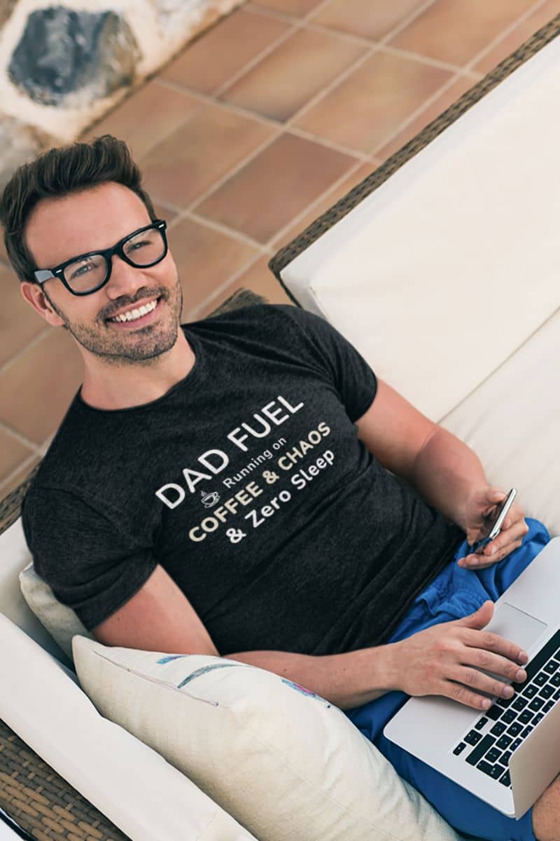 funny t shirts for dads with a man wearing a black t-shirt  featuring the text 'DAD FUEL – Running on Coffee & Chaos & Zero Sleep', sitting on a couch with a laptop.