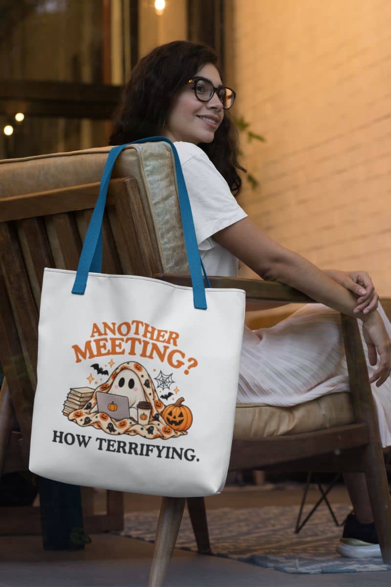 A woman sitting on a wooden chair in a warm indoor setting, beside a funny tote bag with blue handles, featuring a Halloween-themed graphic with the text 'Another Meeting? How Terrifying.'