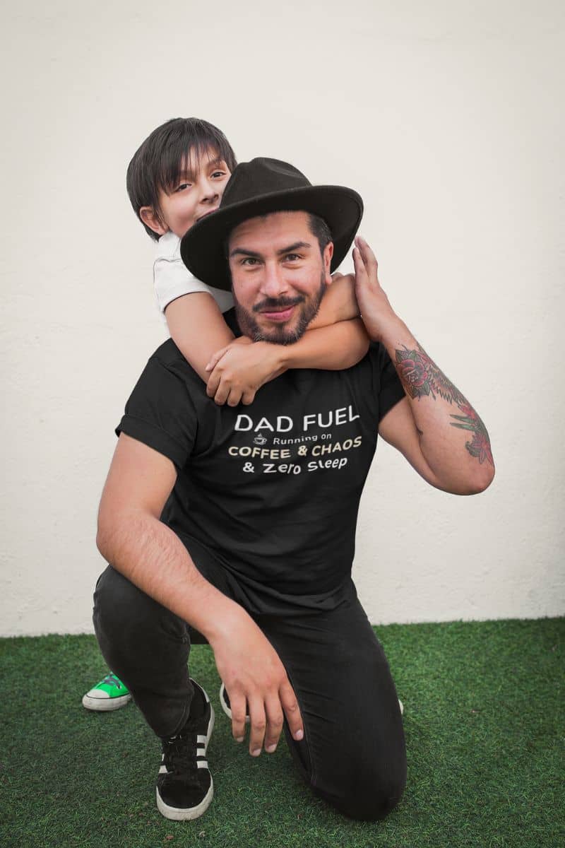 funny tshirt for dad with a man wearing a black t-shirt with 'DAD FUEL – Running on Coffee & Chaos & Zero Sleep' text, holding a child on his back against a white background.