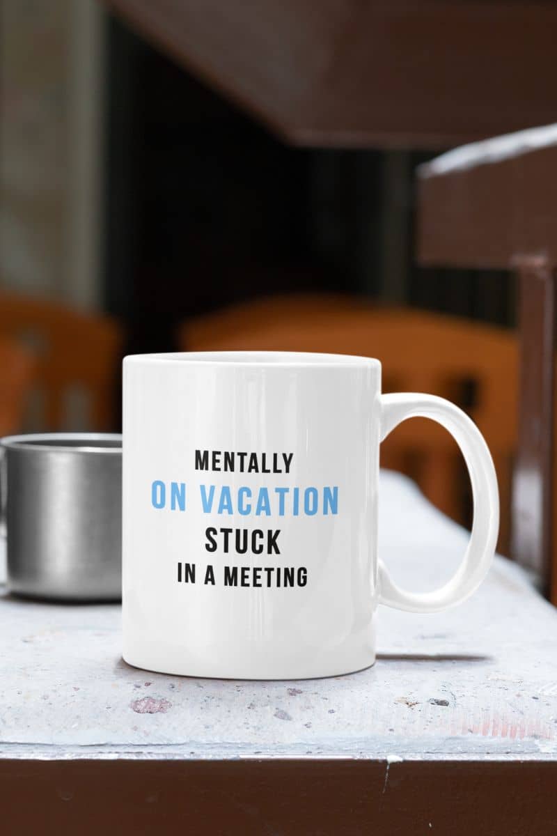 funny vacation mug featuring a white mug with text 'Mentally on vacation, stuck in a meeting' on a table.