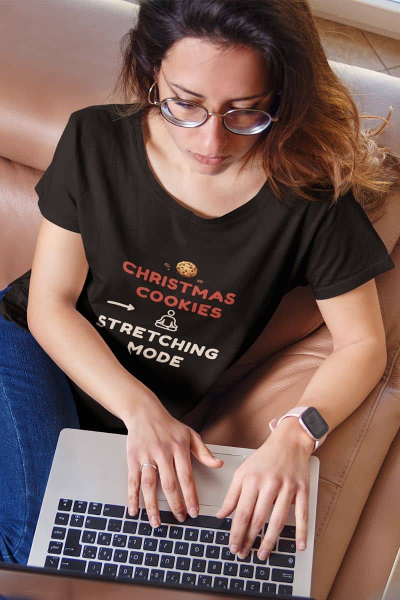 funny wellness t-shirt with a woman wearing a black t-shirt with text 'Christmas Cookies, Stretching Mode', using a laptop on a couch.
