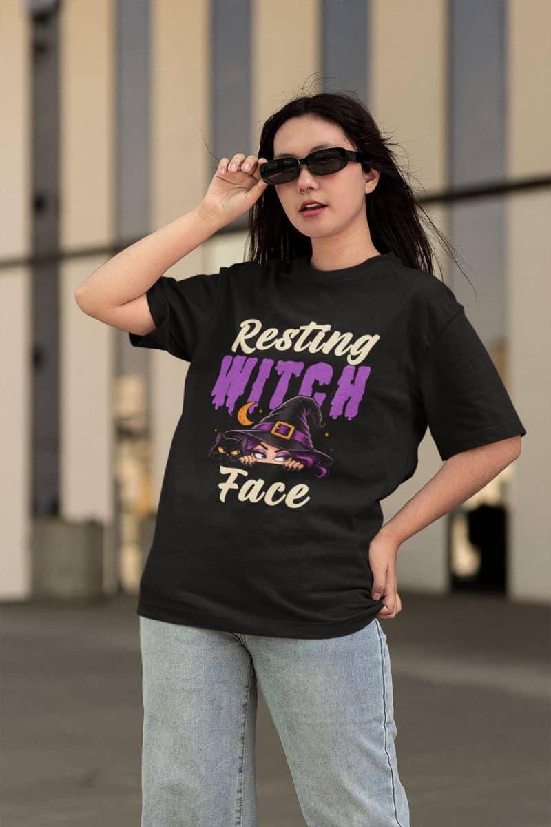 funny witch shirt with a woman wearing a black graphic t-shirt with the text 'Resting WITCH Face' in purple and cream, paired with light-wash jeans, standing outdoors against a blurred building background.