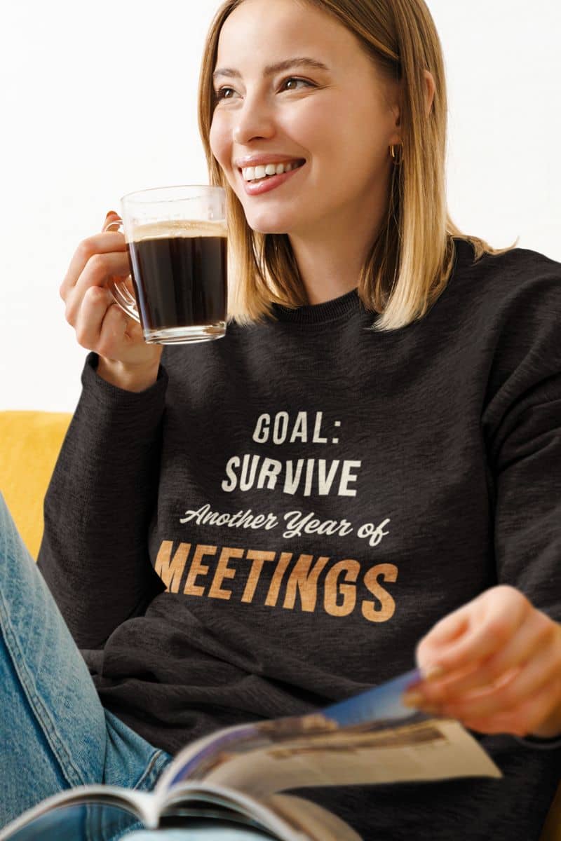 funny work hoodie with a woman wearing a black sweatshirt with text 'Goal: Survive Another Year of Meetings', holding a coffee cup and reading a magazine.