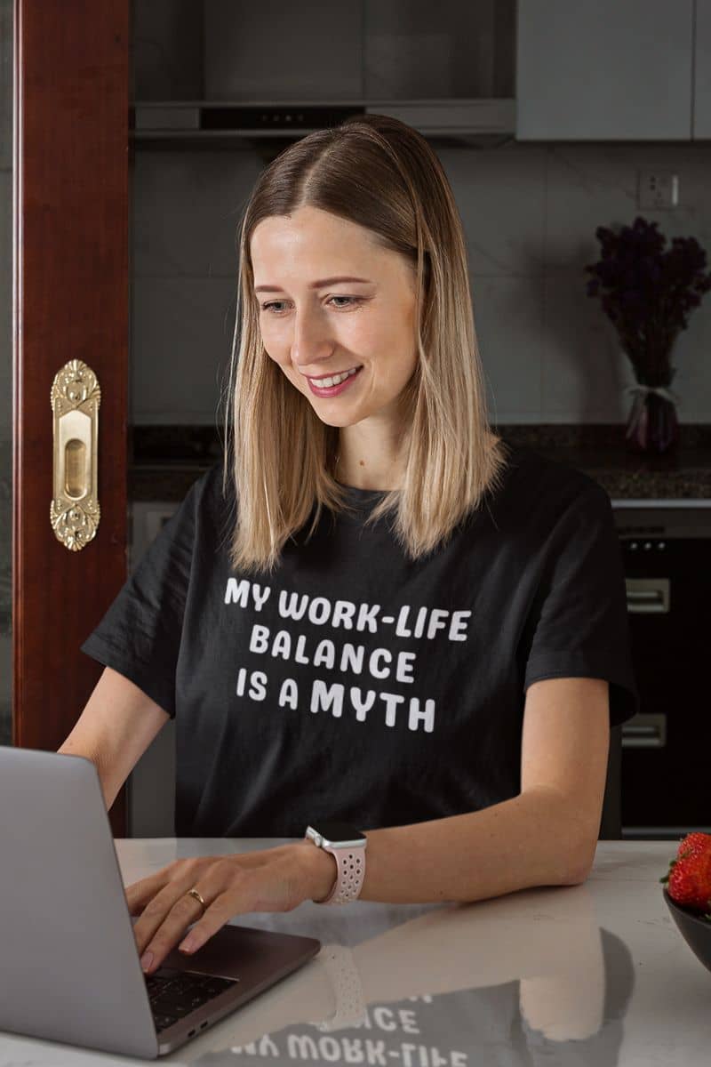 funny work quote t-shirt with a woman wearing a black t-shirt with 'My work-life balance is a myth' text, using a laptop in a kitchen.