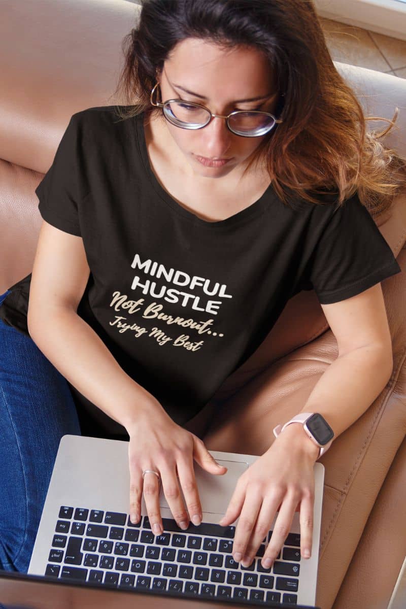 funny work shirt with a woman wearing a black t-shirt with text 'Mindful Hustle – Not Burnout… Trying My Best', using a laptop.