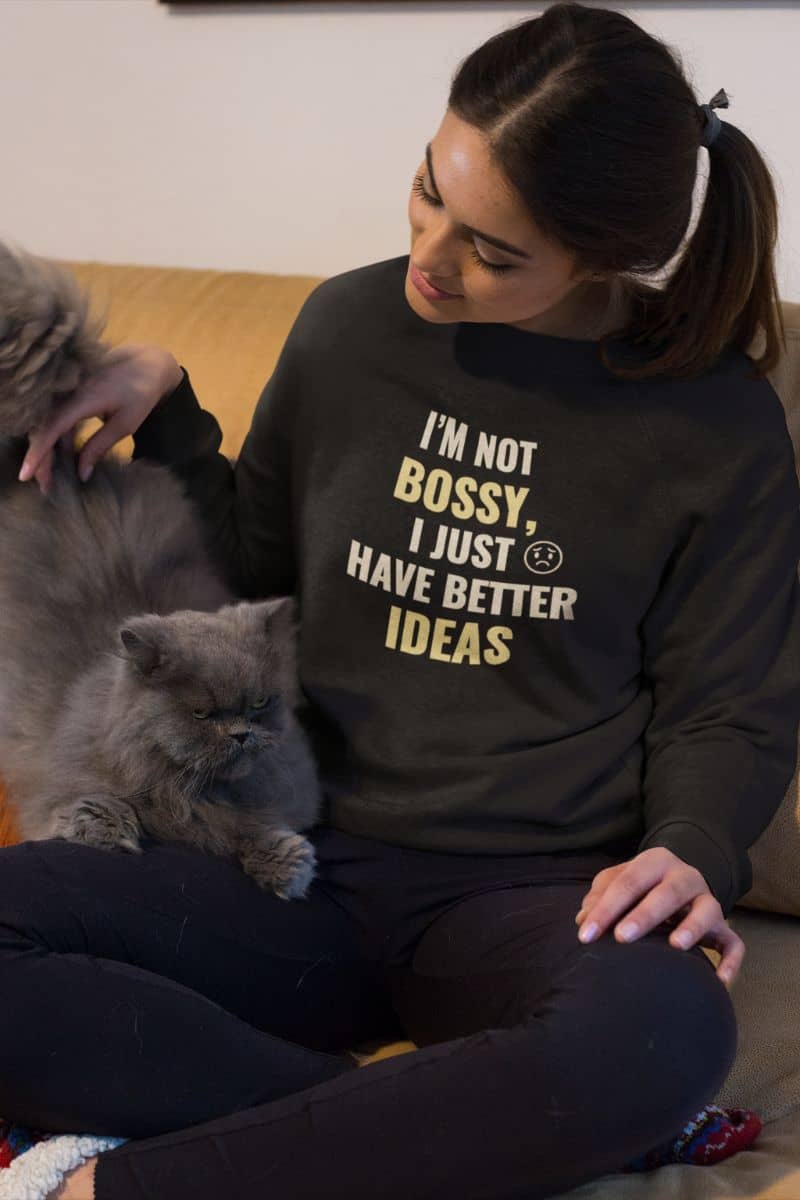 funny work sweater with a woman wearing a black sweatshirt with text 'I’m Not Bossy, I Just Have Better Ideas' sitting on a couch with two cats.