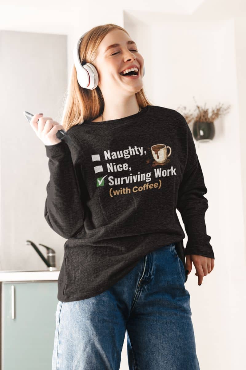 funny work sweatshirt with a person wearing a sweater with text 'Naughty, Nice, Surviving Work (with Coffee)' and headphones, standing in a kitchen.
