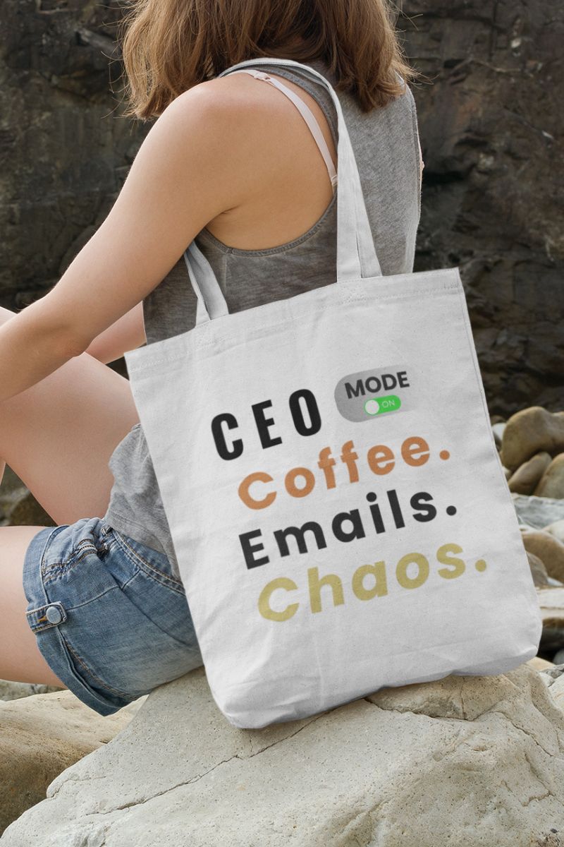 funny work tote bag with a girl holding a tote bag with text 'CEO Mode: Coffee. Emails. Chaos.', sitting on the rock