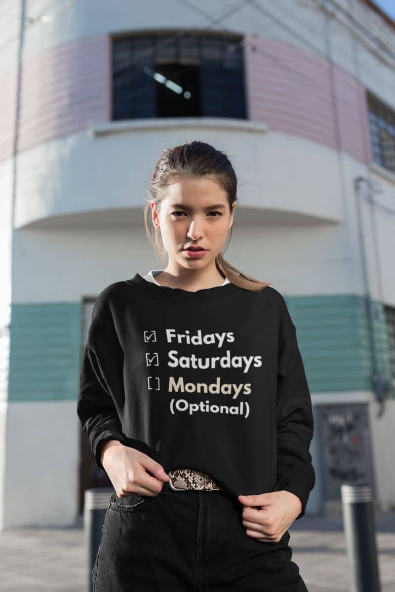 funny workweek sweatshirt with a woman wearing a black sweatshirt with text 'Fridays and Saturdays, Mondays (Optional)' standing in front of a building.