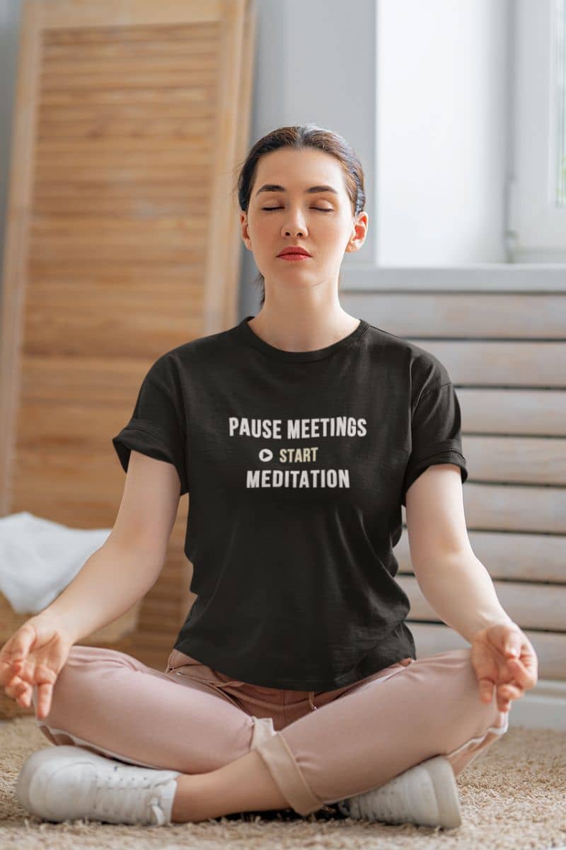 Funny zen t-shirt featuring a woman wearing a black t-shirt with the text “Pause Meetings, Start Meditation” while meditating and doing laundry.