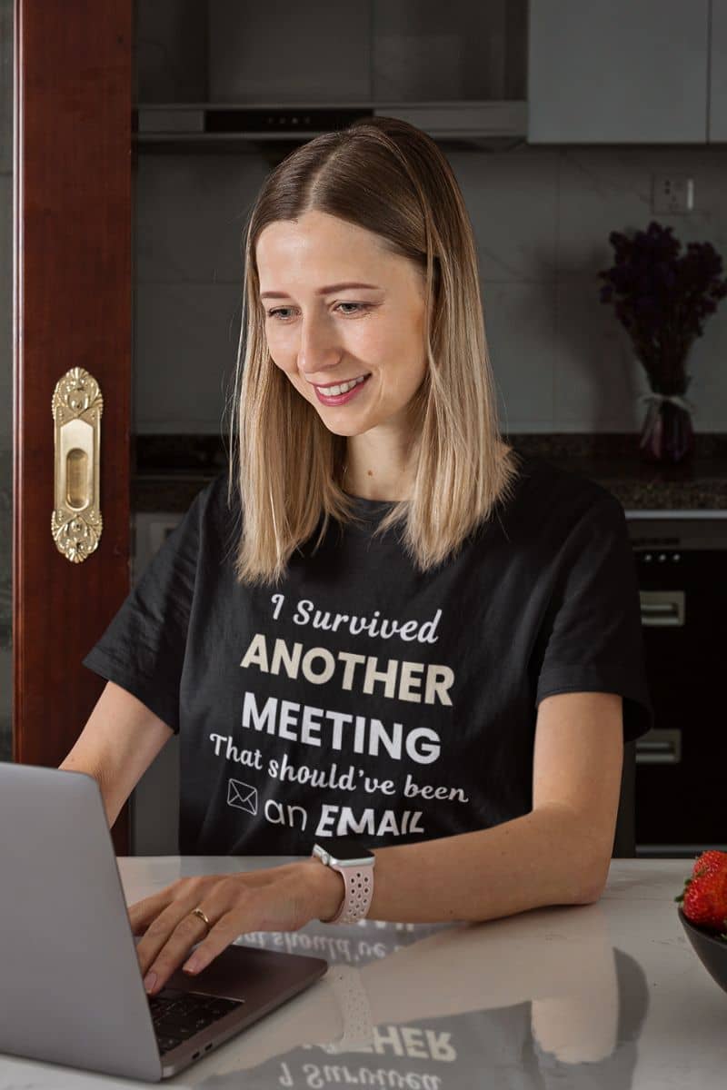 funny zoom t-shirt with a woman wearing a black t-shirt with text 'I Survived Another Meeting That Should’ve Been an Email', sitting at a table with a laptop in a kitchen.